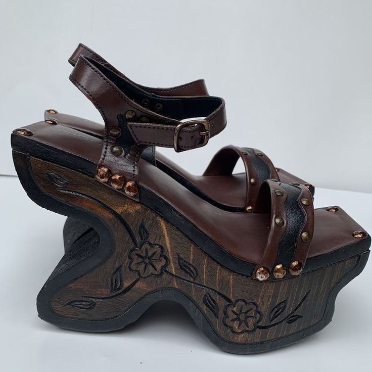 the image shows a pair of brown leather platform sandals with wooden wedges. the wedges are carved with floral designs and have black soles. the sandals have brown leather straps with buckles and are adorned with rivets.
