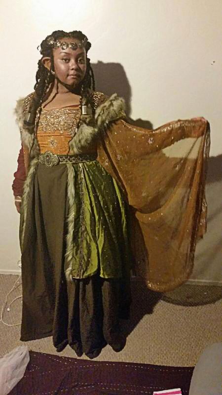 a young woman in an elaborate fantasy costume stands indoors against a neutralcolored wall.  she appears to be cosplaying as an elf or similar fantasy character. her costume features a long dark brown dress layered with a olivegreen velvet overskirt and a flowing, sheer gold shawl.  she wears a furtrimmed cloak, a golden headpiece or tiara, and has long dark braids. the setting is simple, with a visible portion of carpeting on the floor.