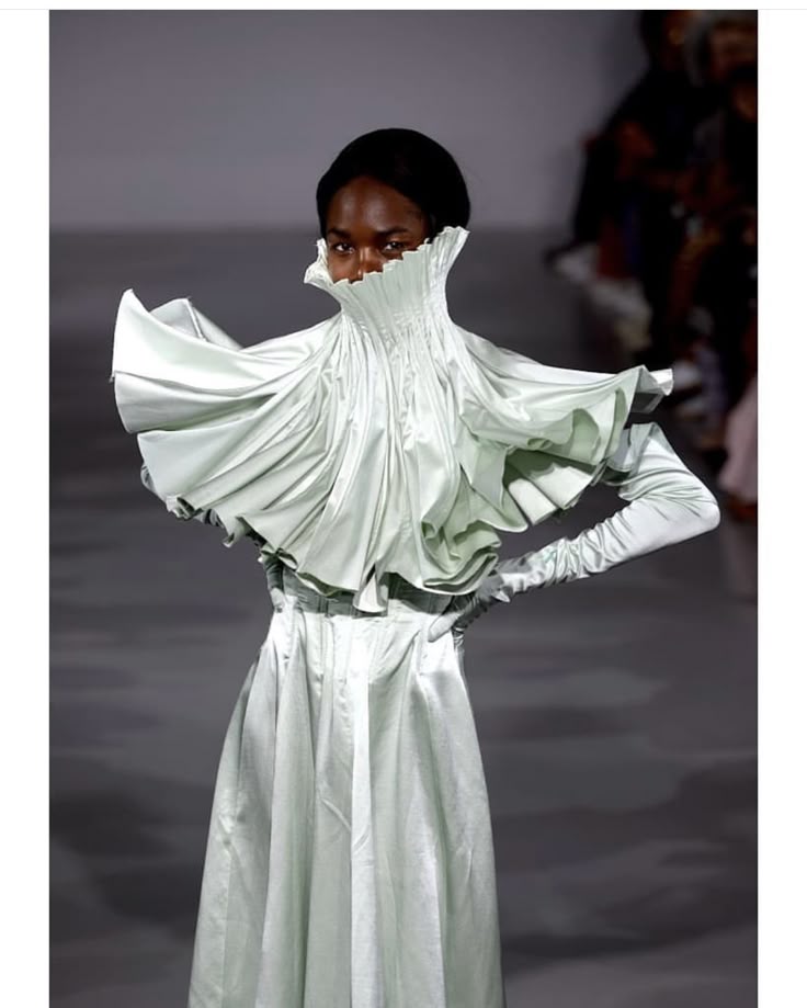 a model poses on a runway in a pale green satin dress with a high, ruffled collar and long gloves. she is partially obscured by the collar, with only her eyes visible. the dress is flowing and dramatic, and the setting is a fashion show. there are other people in the background.