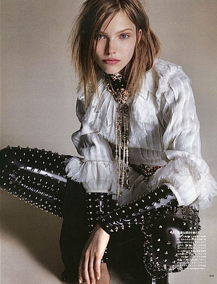 a woman poses for a fashion portrait. she is wearing a white, ruffled shirt with a long, decorative necklace and black, studded pants. she has blonde hair and blue eyes, and she is looking directly at the camera. she is sitting in a crouched position.