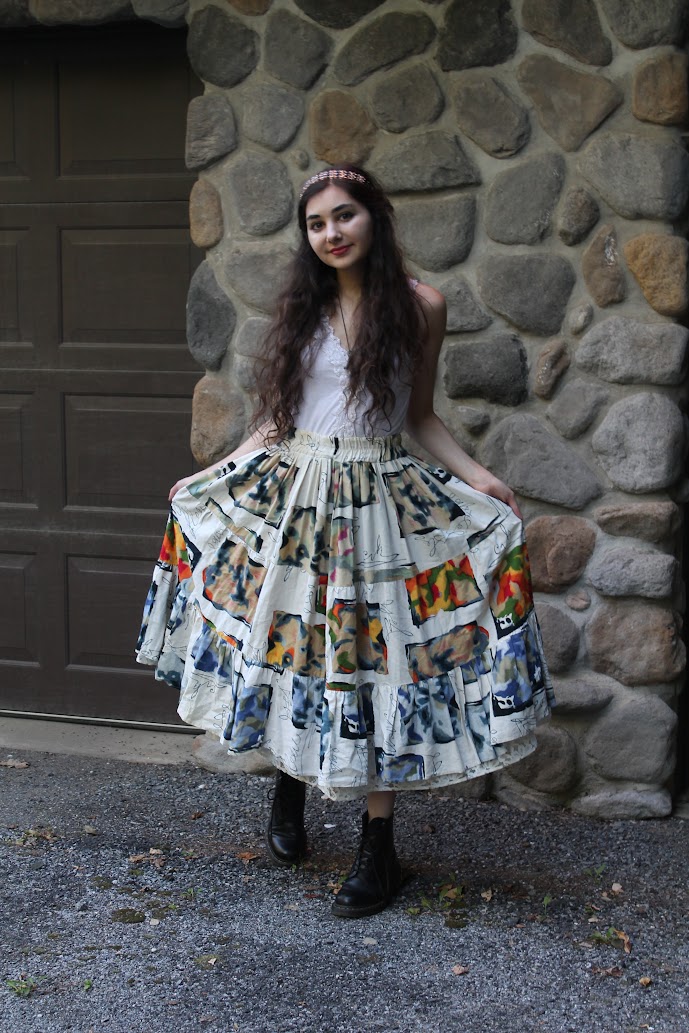 a young woman with long brown wavy hair wears a vintagestyle patchwork maxi skirt and a white camisole top. she accessorizes with a headband and black doc martens. the photo is taken outdoors, with the girl posing against a stone wall.