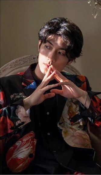 a young man with styled black hair sits in a relaxed pose, his hands gently clasped in front of his face. he wears a black shirt and a vibrant, patterned jacket with splashes of red, white, and blue. the background is softly lit, creating a gentle contrast with the more illuminated subject. the overall mood is contemplative and stylish.
