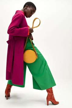 a woman is wearing a long magenta coat with a green wideleg pant and red boots. she's carrying a bright yellow round bag. she's standing in a studio against a white backdrop.