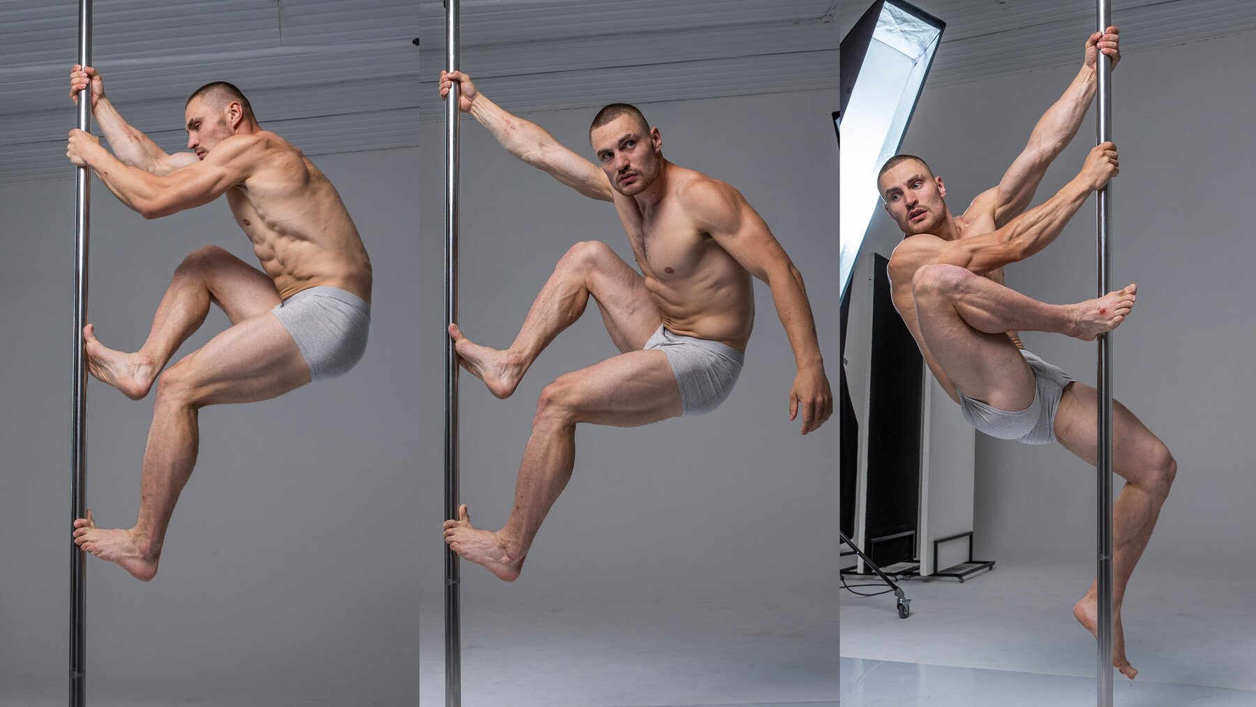 a man is dancing on a pole in a studio. he is shirtless and wearing grey underwear. the background is white and there is a reflection of him in the background. he is muscular and toned. he is posing and dancing on the pole. the light is bright and there are shadows in the background. this is a photo of a man dancing on a pole.