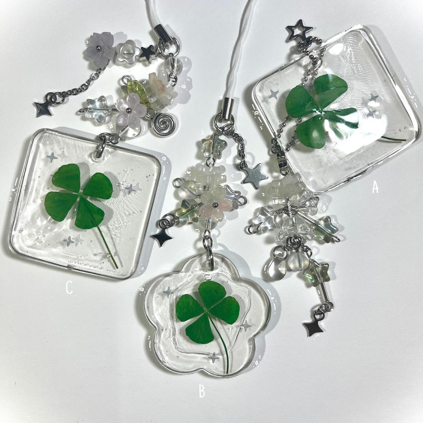 the image shows three different resin keychains, each featuring a fourleaf clover encased within. they have different shapes: square, flower and square. each keychain has additional charms attached, including flowers, beads, and stars. they are arranged against a white background.