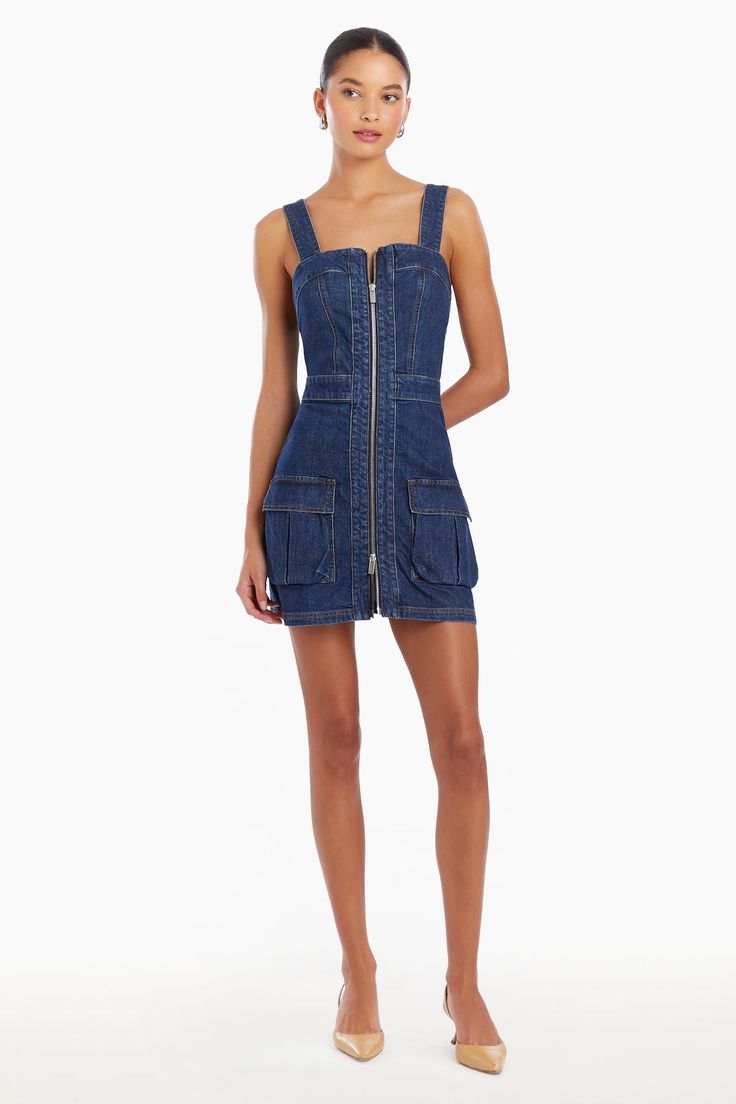 a woman standing in a white setting, wearing a denim mini dress with a zipper down the front and pockets on the sides. she is wearing nude heels.