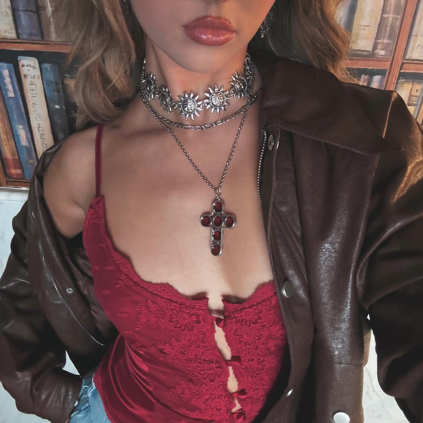 a woman is wearing a red lacetrimmed top with a brown leather jacket draped over her shoulders. she is adorned with multiple silver necklaces, including a sunburst choker and a long chain with a large cross pendant containing red stones. behind her is a blurred bookshelf.