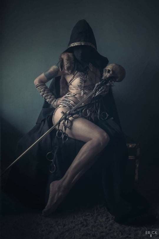 a woman in a dark hooded cloak sits and poses with a skull in her hand, holding it in a mysterious and gothic manner. the woman's pose is sensual and powerful, and she is surrounded by a dark and shadowy atmosphere. this image is full of symbolism and evokes a sense of mystery and intrigue.  the woman is a witch or sorceress.  there is a sense of foreboding, but she seems strong and powerful.