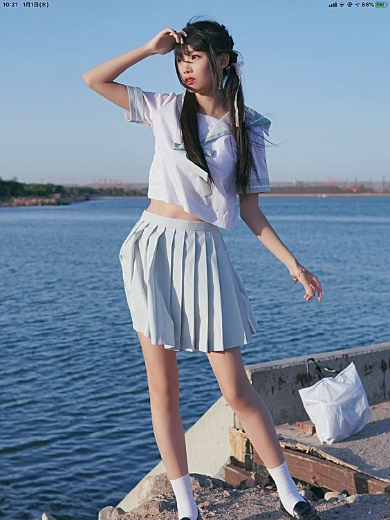 a young woman in a sailorstyle crop top and pleated skirt stands on a dock by the ocean. she is wearing white socks and black loafers, and has her hair in pigtails. the background features a clear blue sky and the ocean, with a glimpse of the horizon in the distance. the image is brightly lit with natural sunlight, giving it a summery feel.