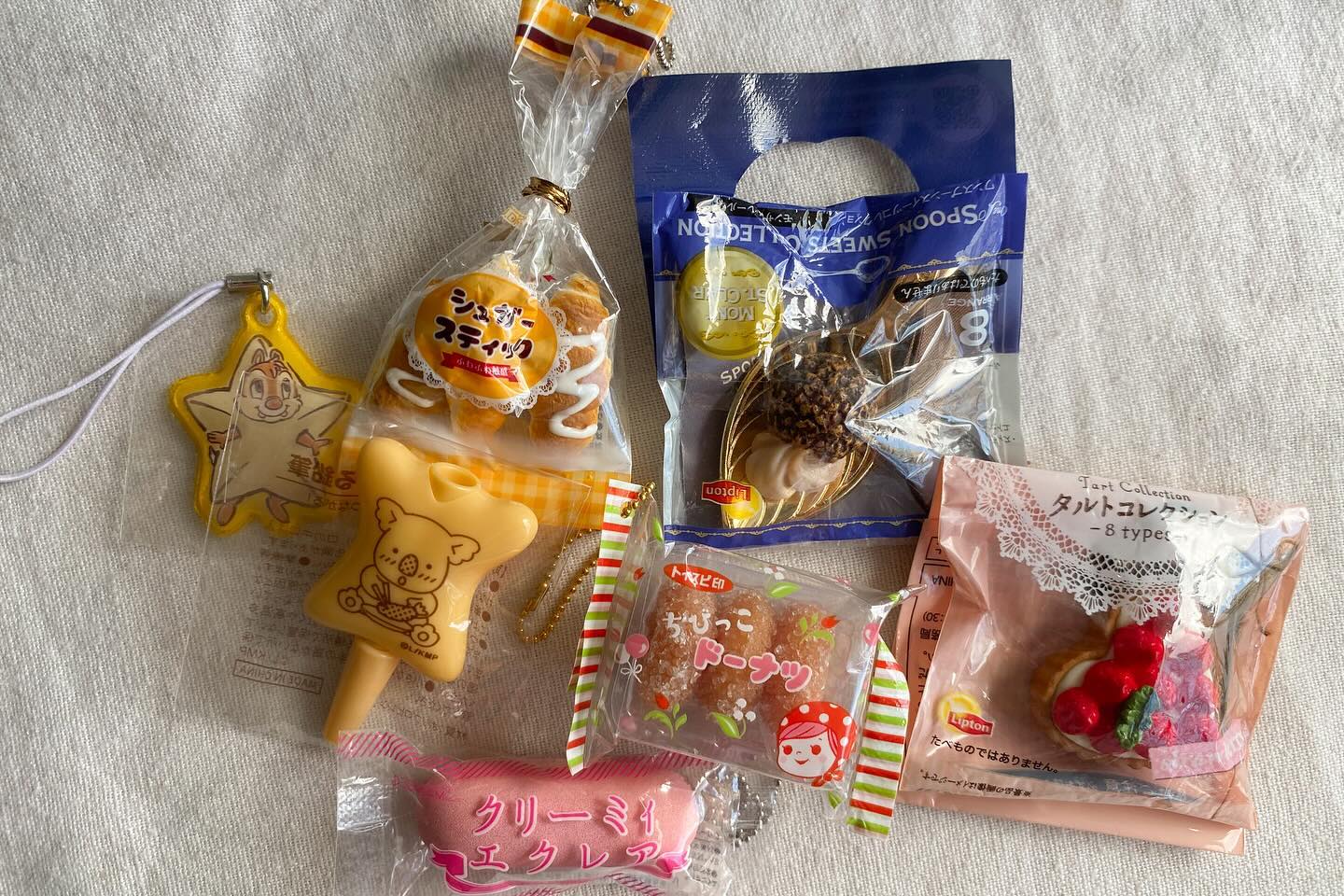 the image shows a collection of packaged food items, including desserts and snacks. there are packaged sugar sticks, donuts, an eclair, and tarts, some of which are from a "spoon sweets collection" by lipton. there are also two charms or figurines in the image, one depicting chip from chip and dale and another of a yellow koala bear. the food is all packaged in plastic wrapping.