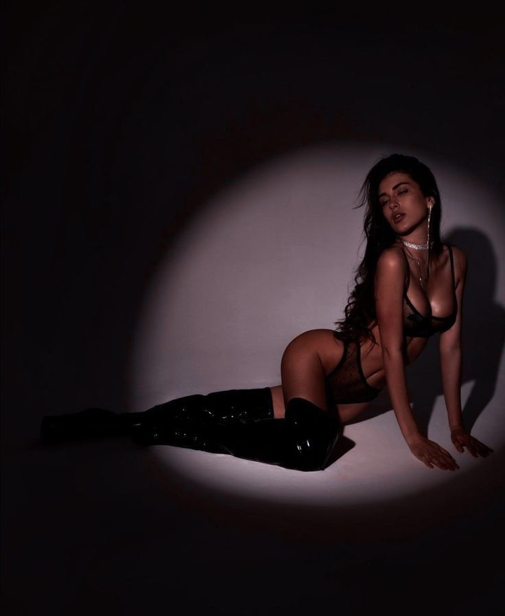 the image is a stylized studio shot of a sensual young woman.  she is reclining on the floor, wearing black lingerie (a lacy bra and panties) and long black leather thighhigh boots. her long dark hair is loose, and she has on minimal makeup and delicate jewelry.  a single spotlight illuminates her against a dark, plain background, creating a dramatic and glamorous effect. the overall mood is suggestive and alluring.