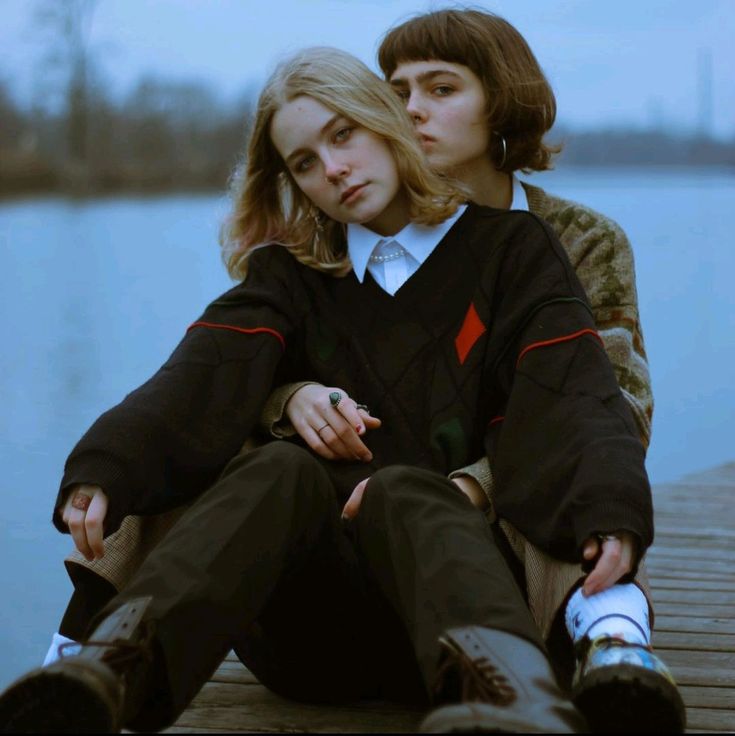 two young women, dressed in trendy clothing, sit on a wooden dock by a lake, smiling and looking at the camera. the girl on the left is wearing a black sweater with a red diamond pattern, while the girl on the right is wearing a brown and white sweater. both girls are wearing dark pants and black boots. they are posed closely together, with their arms around each other, showing a sense of intimacy and connection. they appear to be best friends, enjoying each other's company and creating a sweet and loving atmosphere.