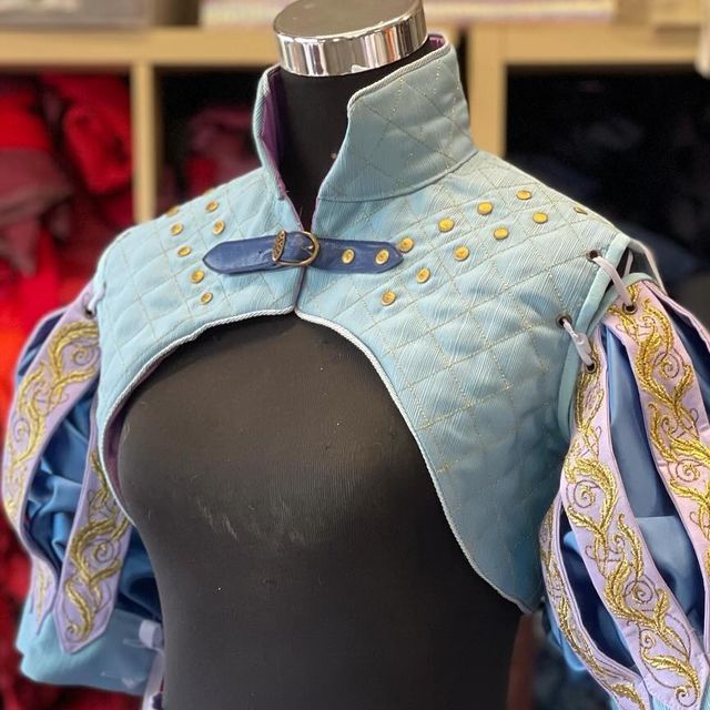 a cropped image features a light blue, quilted bolero with lavender sleeves and gold trim displayed on a black mannequin. the bolero has a high collar and is fastened at the front with a blue buckle. small gold studs are arranged in a pattern on the quilted material. the sleeves are puffed and gathered, with intricate gold embroidery. the bolero and sleeves are displayed against a backdrop of shelves with various items.