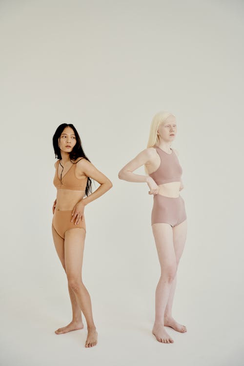 two women, one with dark hair and the other with light blonde hair, stand sidebyside in a minimalist studio setting. they are both wearing simple, comfortablelooking lingerie sets in nude and pale pink colors. the image highlights body diversity and inclusivity.  the background is plain white, allowing the focus to stay on the models.