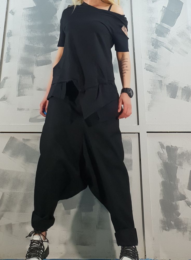 a woman wearing a stylish black outfit. she is wearing loose fitting, baggy black pants and a black, off the shoulder top that is tied at the waist, with a few strings hanging loosely, which gives it a  casual, cool and trendy look. she is posing for a photo while standing against a white wall.