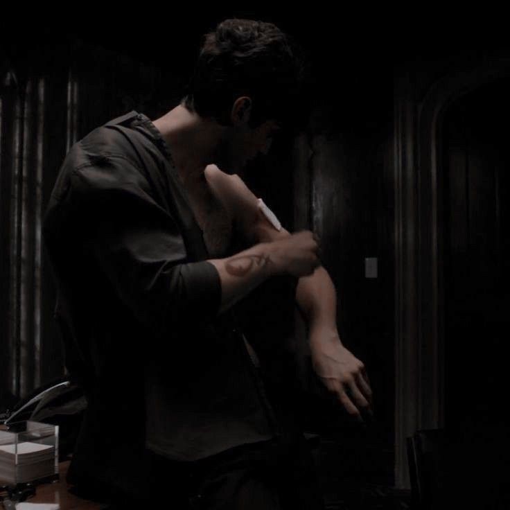 a dimly lit image shows a man from the side, leaning over with his head slightly bowed. he is wearing a dark, loosefitting shirt or jacket and has a tattoo on his upper arm. he appears to be tending to an injury on his arm with a bandage. the scene is dark and shadowy, with only some light illuminating the man and the immediate area around him. the background includes dark walls, a door, and some furniture.
