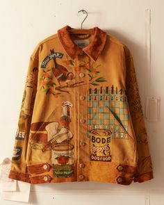 a light brown suede jacket hangs on a rack. it is painted with a variety of images and patterns, such as birds, coffee, checkers, and art.