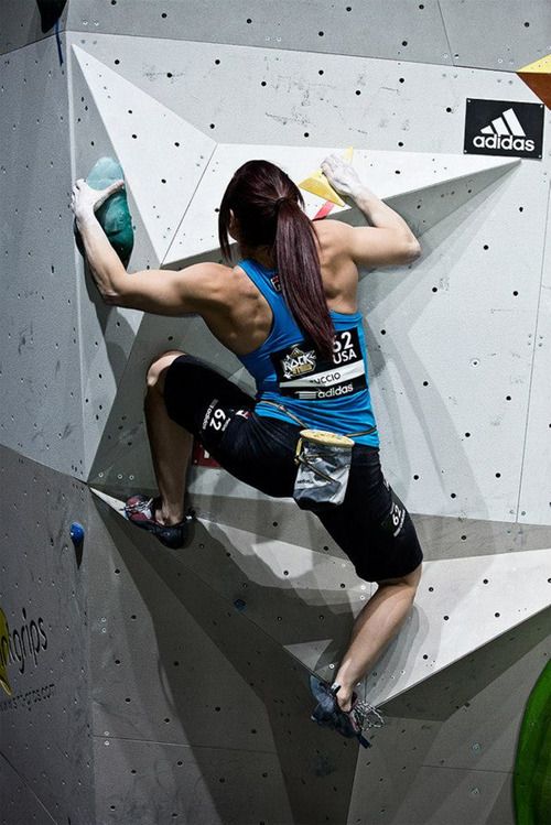 a woman wearing a blue tank top and black pants is rock climbing on an indoor climbing wall. she is climbing a difficult route and looks focused and determined. she is wearing climbing shoes and has her hands on the wall. the wall is white and has many holds. she is wearing an adidas logo.