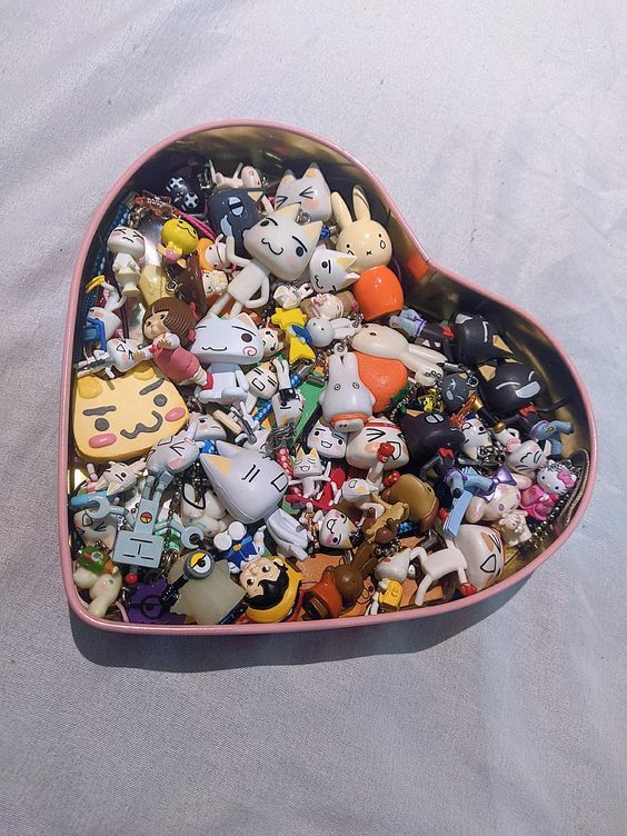 a heartshaped tin filled with a collection of toy figurines and keychains. the items include various cartoon characters, cats, rabbits, and other small items, forming a diverse assortment of miniature collectibles. the tin has a pink trim.