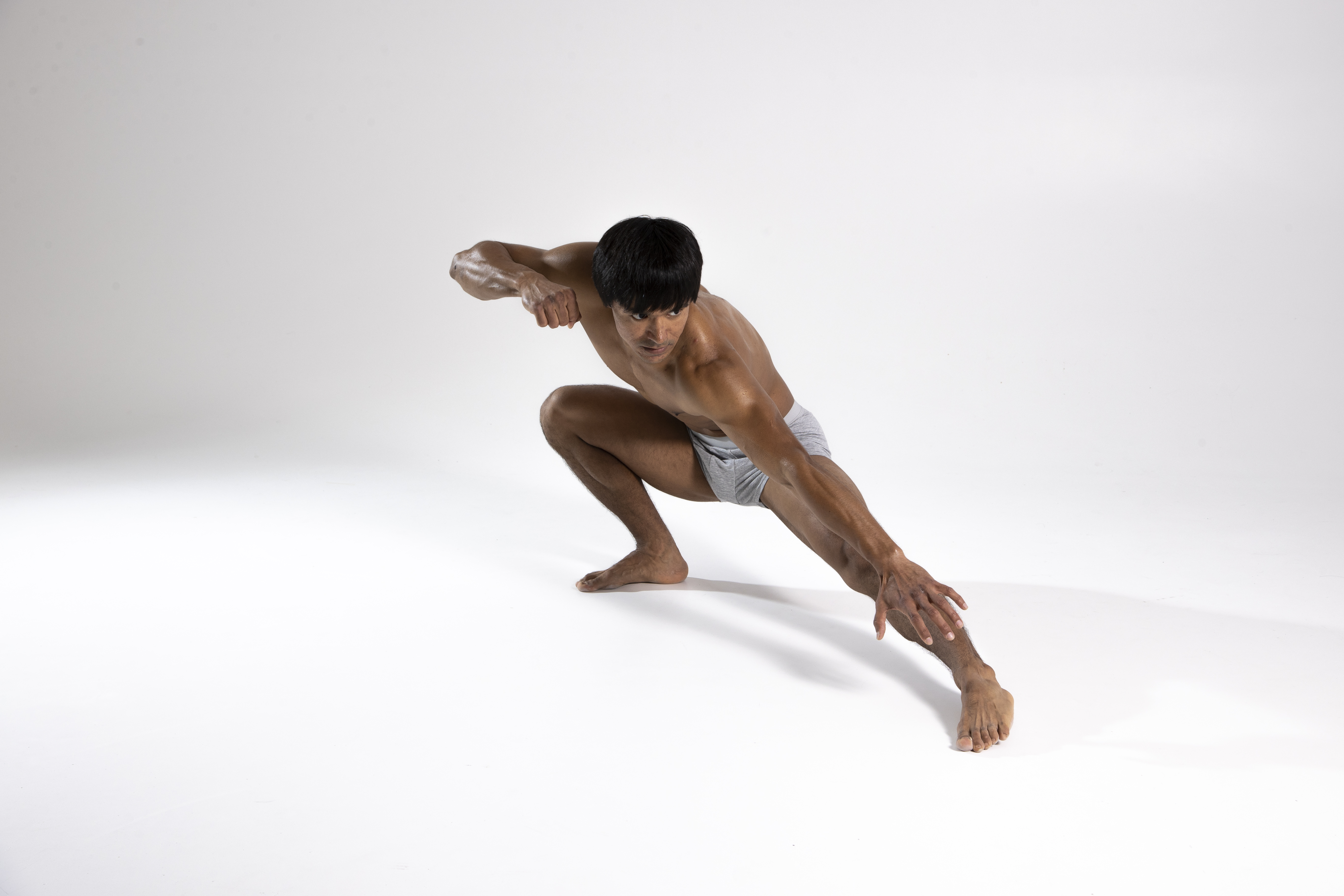 a toned, barefoot man in grey underwear is captured in a dynamic stretching pose against a stark white studio backdrop. he is crouching with one leg extended forward, hand reaching towards the floor, while the other leg is bent beneath him. his body showcases athleticism and a dedication to fitness.