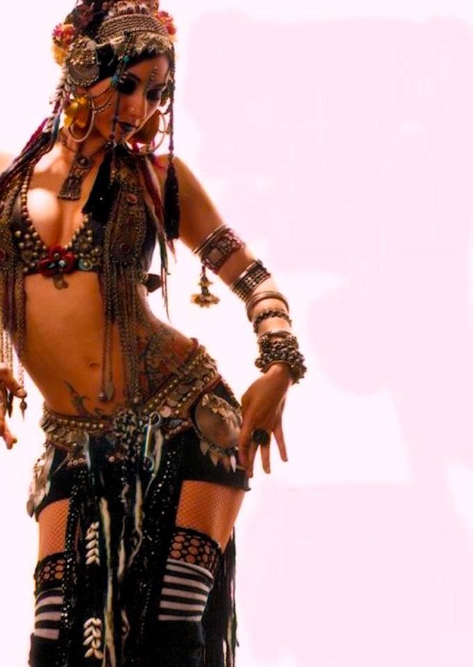 a belly dancer wearing a traditional belly dancing costume with lots of  jewelry is posing in front of a white background. the dancer has on several bracelets and necklaces,  and she has a headscarf and a veil on.