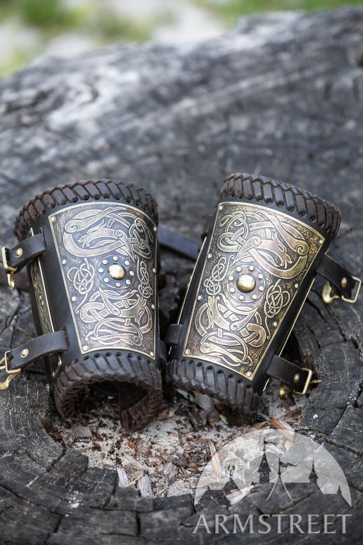 the image shows a pair of leather arm guards with intricate celtic knot designs, placed on a weathered wooden surface. the arm guards are black leather with a goldcolored metal plate adorned with the celtic knot pattern and a central rivet. they feature leather straps and brass buckles for fastening. the background is slightly blurred, suggesting an outdoor setting. the word "armstreet" appears in the bottom right corner of the image.