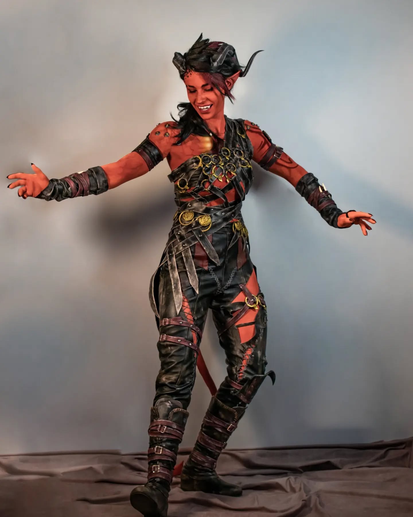 a fullbody shot of a person in elaborate cosplay portraying a tiefling, a type of demon from fantasy settings like dungeons & dragons. the cosplayer has red skin, black horns, and is wearing a meticulously crafted costume that features dark leather armor, intricate straps, and a ripped leather pants design with red accents. the outfit is highly detailed, suggesting significant time and effort in its creation. the cosplayer has a playful and fierce expression, striking a dynamic pose that showcases the costume's details and their confidence in the character portrayal. the background is a plain, light gray, which draws focus to the cosplayer and their costume. the image appears to be professionally photographed in a studio setting.