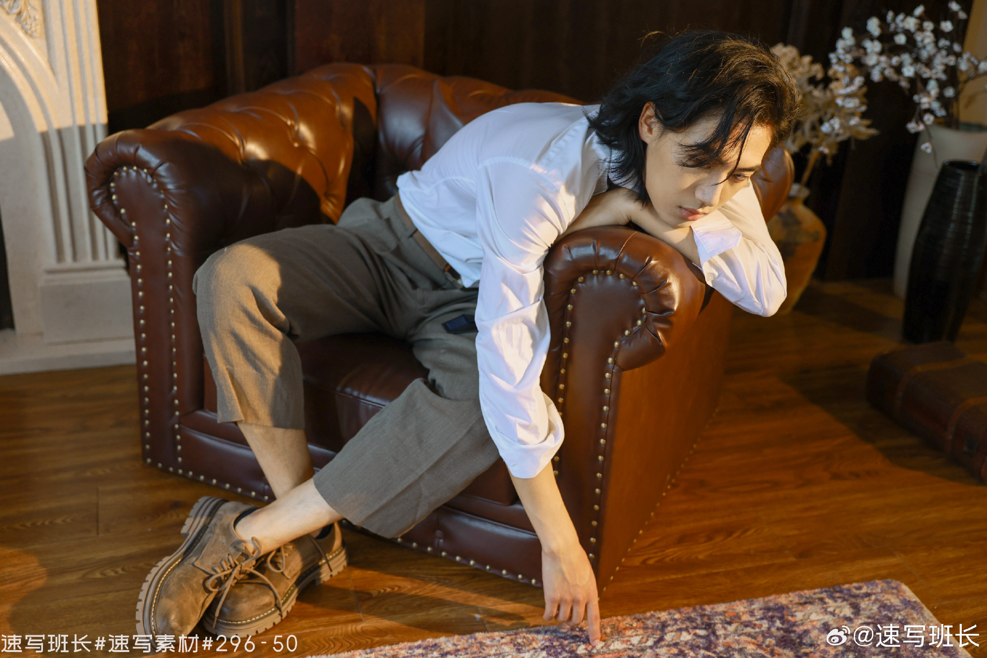 a young man with black hair is posing on a brown leather sofa. he is wearing a white shirt and brown pants, and he is looking down at the floor. the photo is taken in a living room with a fireplace in the background. the lighting is natural and comes from a window offcamera. the man is wearing brown shoes.