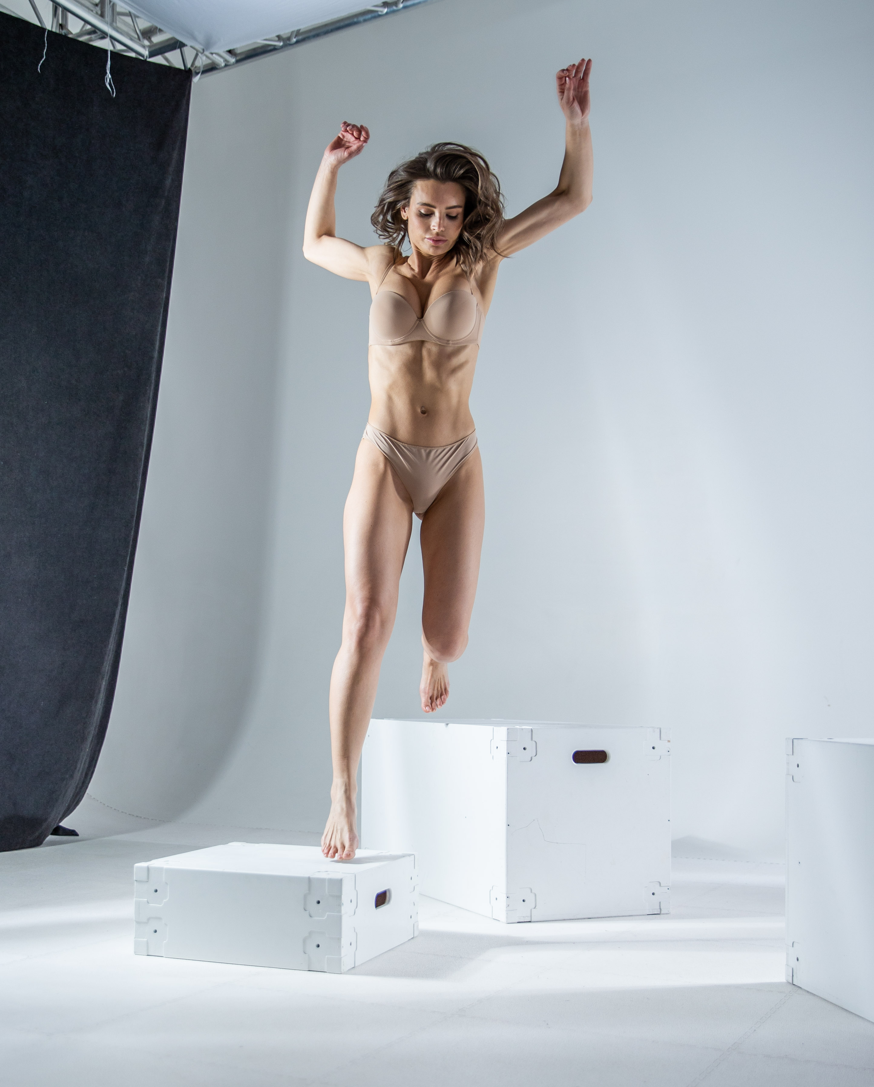 a woman in lingerie is jumping between white boxes in a studio setting. she is midair, with her arms raised.