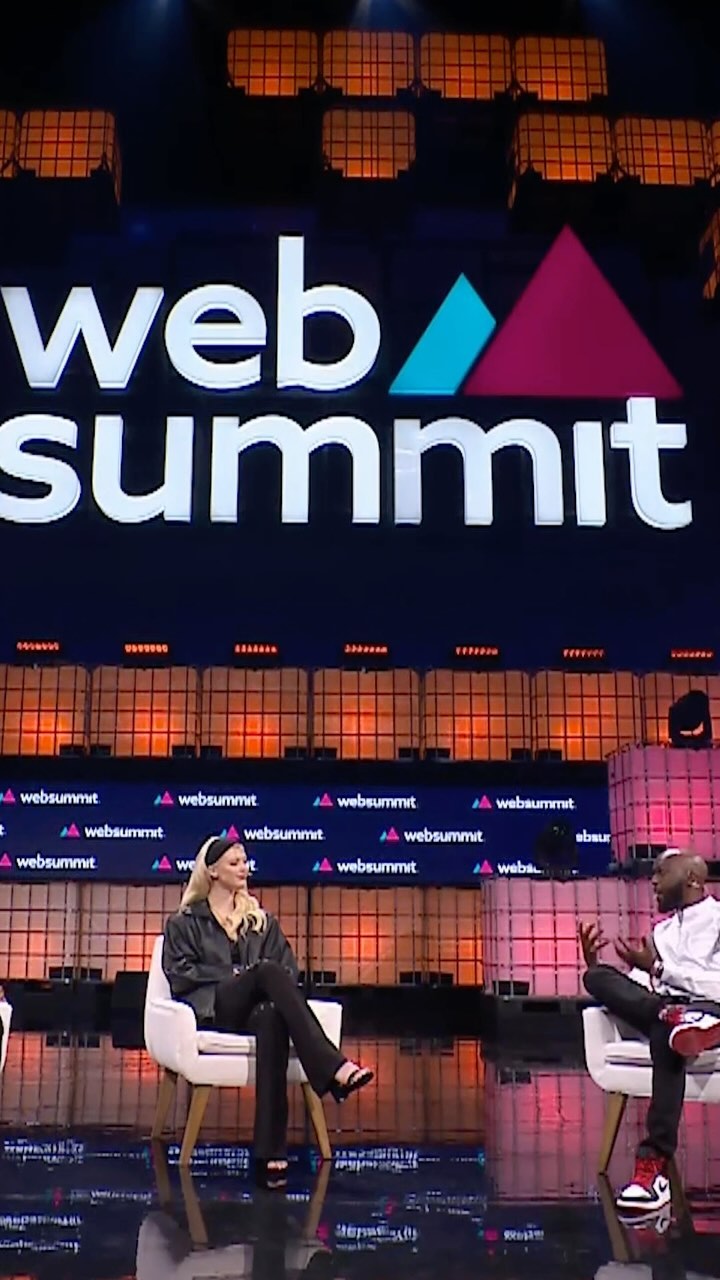 two people, a blonde woman and a black man, are sitting in white chairs on a stage during a web summit conference. the stage has a large screen displaying the "web summit" logo. the woman is wearing a black leather jacket and black pants, while the man is wearing a white shirt, black pants, and red sneakers. they appear to be engaged in a discussion. the background includes a digital display with the "web summit" logo repeated.