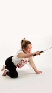 a woman in dark pants and white shirt, stained with fake blood, is captured midcrawl against a white background.  she is reaching out with her right arm, brandishing a sword.  her expression is one of fierce determination. the lighting is soft, suggesting a studio setting.