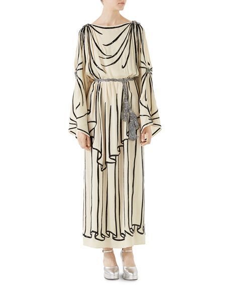 a woman is wearing a long dress in this full shot. the dress is cream colored with black stripes and has a belt with tassels. the dress has a loose, flowing style. she is also wearing silver shoes. the background is plain white.