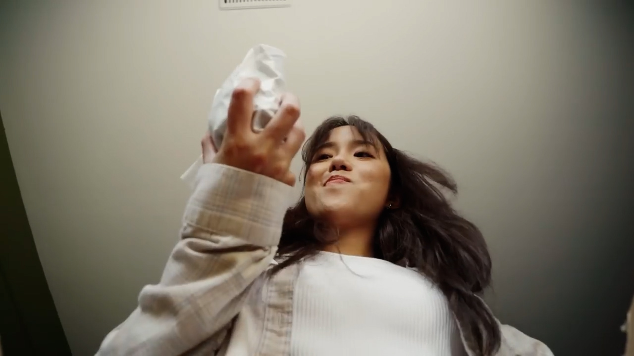 a young woman with long dark hair is captured from a low angle, about to playfully throw a crumpled ball of paper. she is indoors and wears a casual, lightcolored outfit. her expression suggests a lighthearted and mischievous mood.