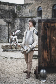 a young man dressed in medieval knight's armor stands in front of a large wooden door in a stone castle courtyard. he is holding a sword and wearing a breastplate and other defensive gear. a second, more experienced knight is visible in the background.  the overall scene is suggestive of a historical reenactment or a period drama.