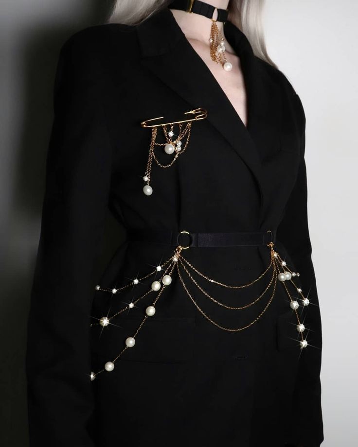 a woman is wearing a black blazer with a black belt and various pearl and gold chain jewelry. the blazer is accessorized with a large gold safety pin brooch adorned with hanging pearl and gold chain embellishments. the belt has matching pearl and gold chain drapes. the woman also wears a black choker with hanging pearls and chains. the background is a light grey.
