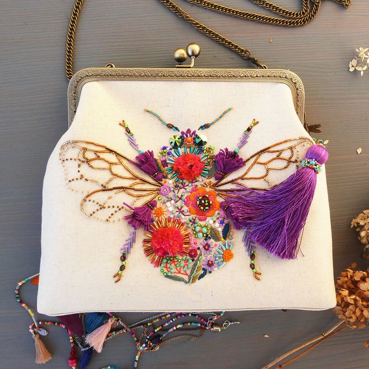 a white vintage clutch purse with a gold chain strap and embroidered with a colorful bee embellished with beads and sequins. the bee is surrounded by flowers and the purse also has a purple tassel. the purse is displayed on a wooden table with a few other beaded necklaces.