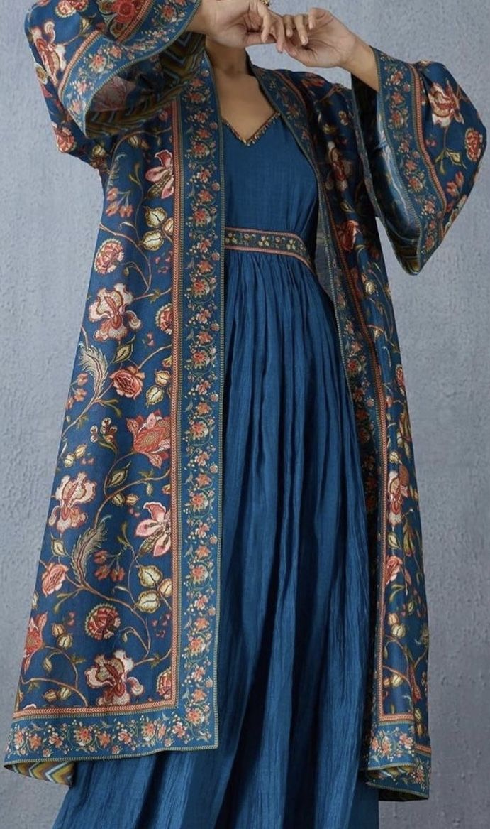 the image shows a person wearing a blue dress with a floralpatterned shawl over it. the dress is long and flowing, with a fitted bodice and a full skirt. the shawl is made of a similar fabric to the dress, with a blue background and a floral pattern in shades of orange, pink, and green. the person is standing with their hands raised to their face, as if they are admiring the dress.