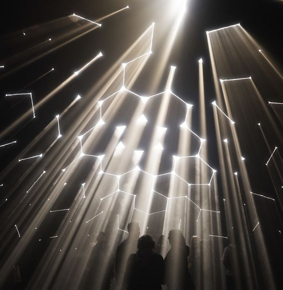 a lowangle shot within a darkened room, illuminated by numerous spotlights directed upwards. the beams of light create a pattern against the ceiling and are scattered with geometric shapes formed by smaller lights. silhouettes of people are faintly visible in the foreground, gazing upward at the display.
