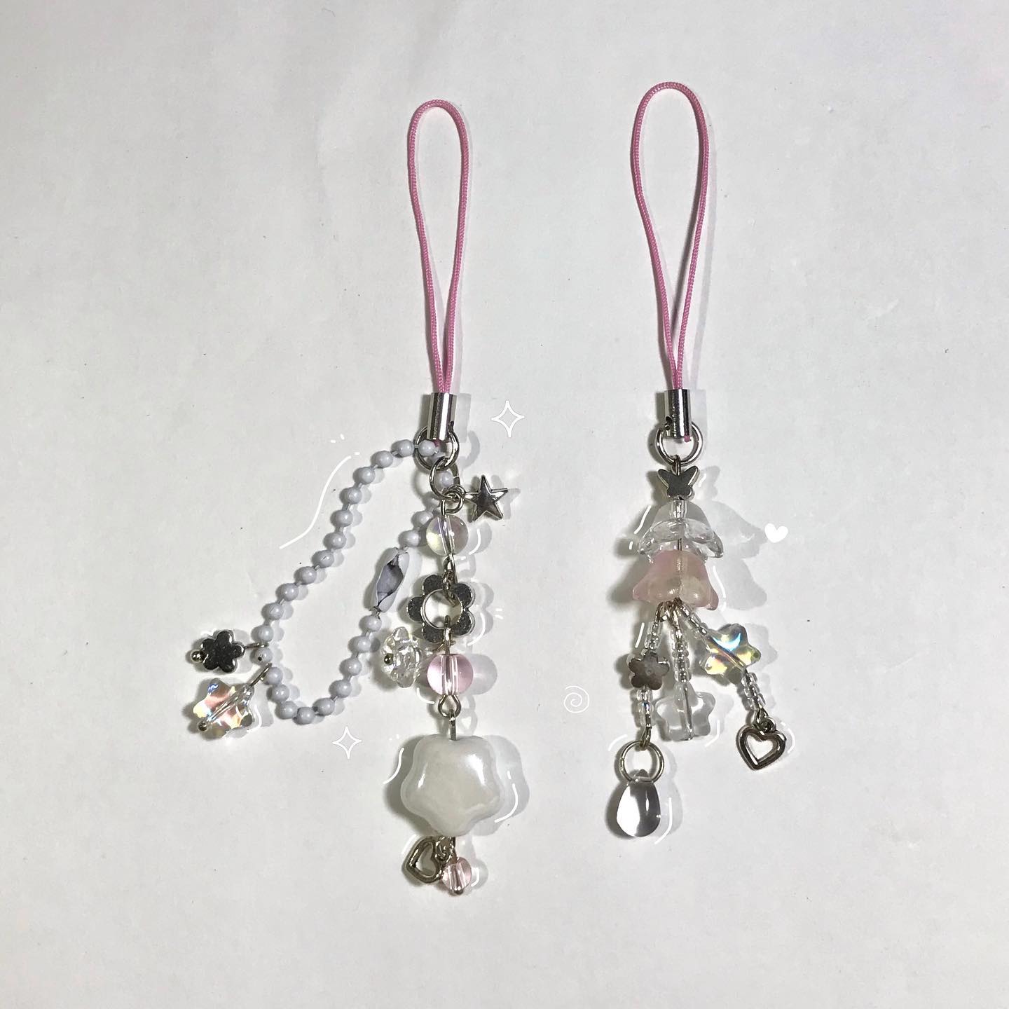 the image features two handmade phone charms with pink straps and various beads and charms in a cute and kawaii style. one charm has a beaded chain, star charm, flower charm, pink and clear beads, and a white flowershaped charm. the other charm features clear and pink shaped beads, heart charm, and other smaller beads.