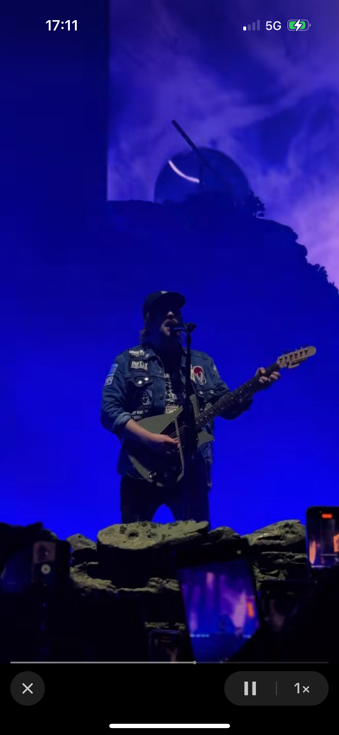 a man with a microphone and a guitar plays a song on stage at a concert. the stage is dimly lit and the background is dark. there is a crowd of people in the background.