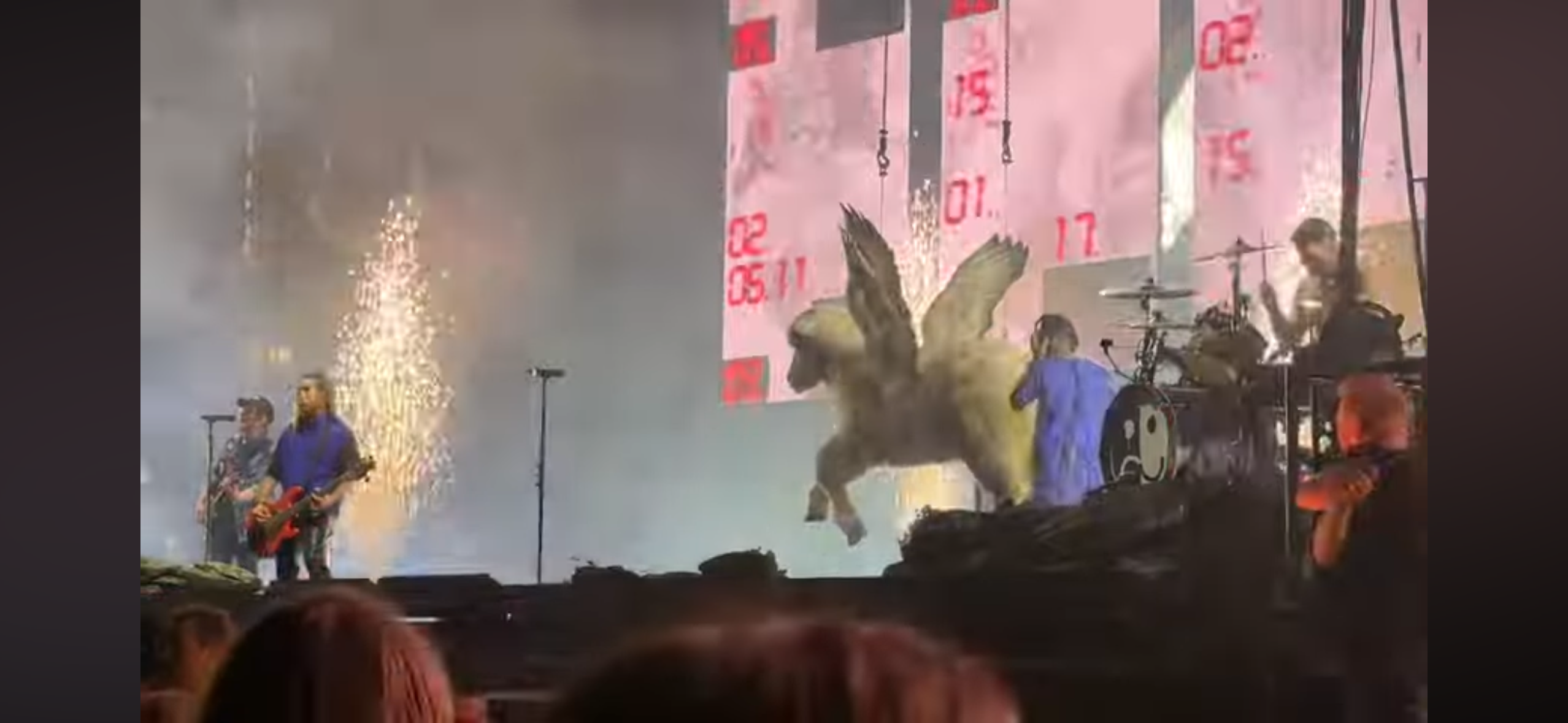 a live band performs on a stage in front of a large crowd of people. fireworks are going off in the background, and there is a large screen behind the band showing a graphic of a winged horse. the band is made up of a singer, guitarist, and drummer. they are all wearing casual clothes. the crowd is cheering and having a great time. the energy is high, and the overall atmosphere is one of excitement and fun.