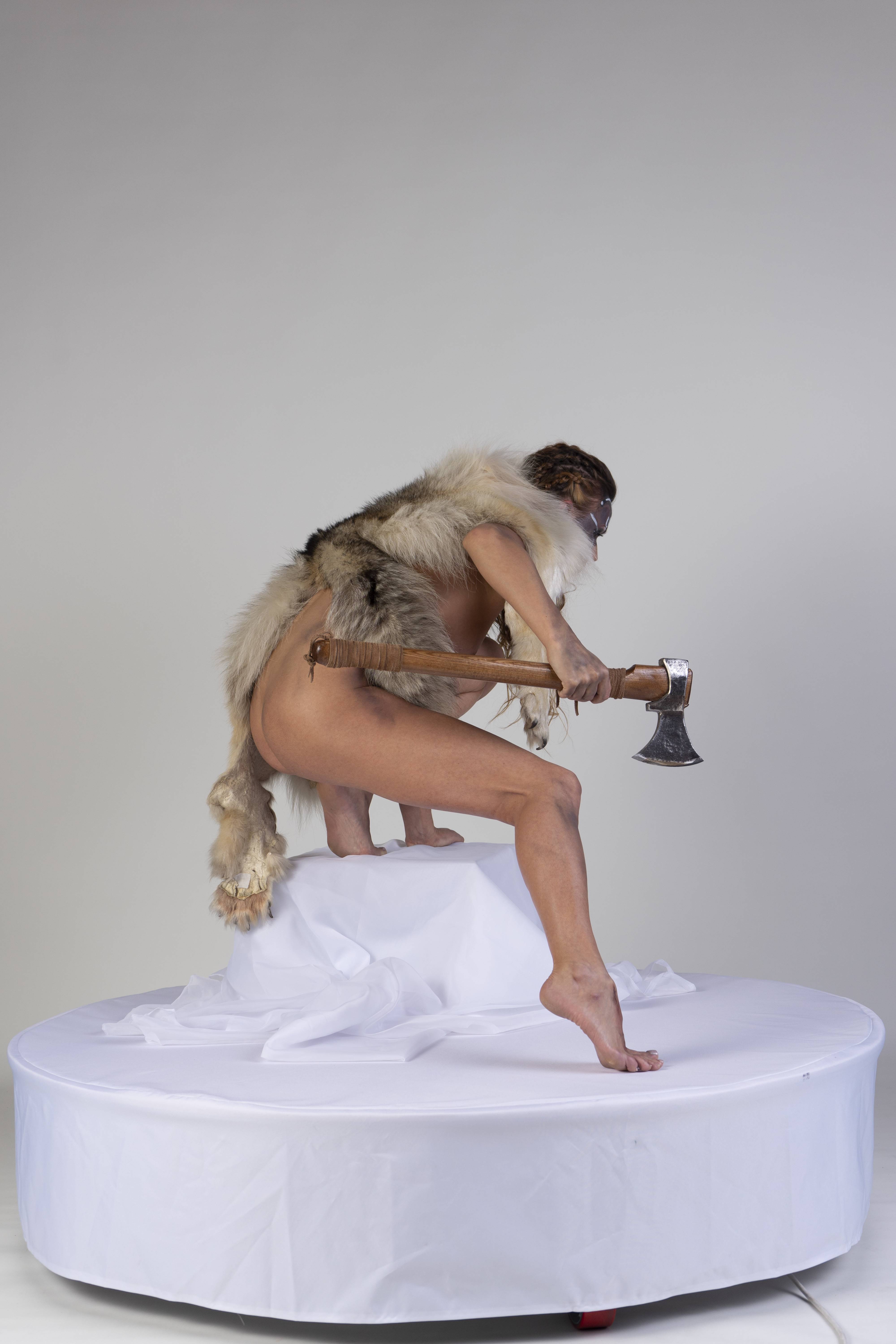 a nude woman stands on a white platform with an axe and wearing a fur pelt.