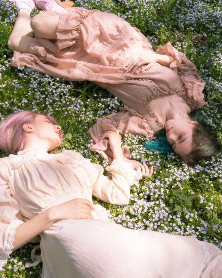 two women are lying down in a field of small white flowers. the woman on the left has pink hair and is wearing a light pink dress with lace trim. the woman on the right has darker hair with a teal tint and is wearing a similar dress with ruffles. they both appear to be sleeping or resting peacefully in a dreamy, serene setting.