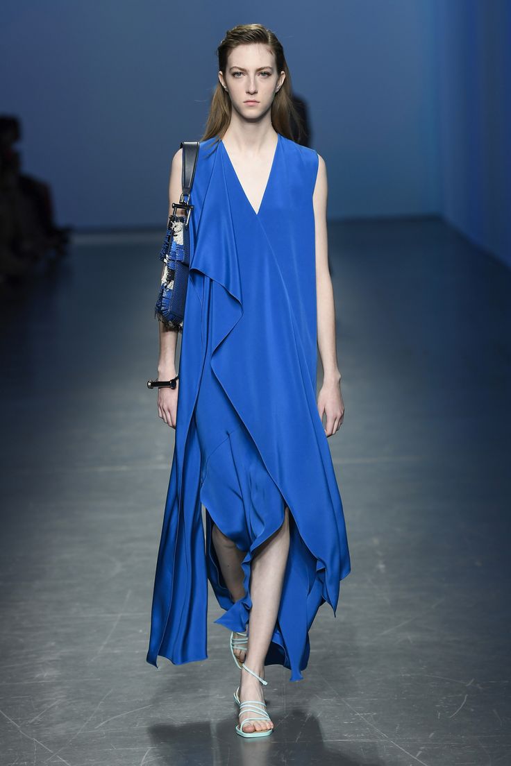 a female model walks the runway in a stunning cobalt blue maxi dress. the dress features a vneckline, asymmetrical draping, and a high slit.  she is carrying a patterned shoulder bag and wearing light blue sandals. the background is a simple, neutraltoned runway. the overall image showcases elegant and sophisticated fashion.