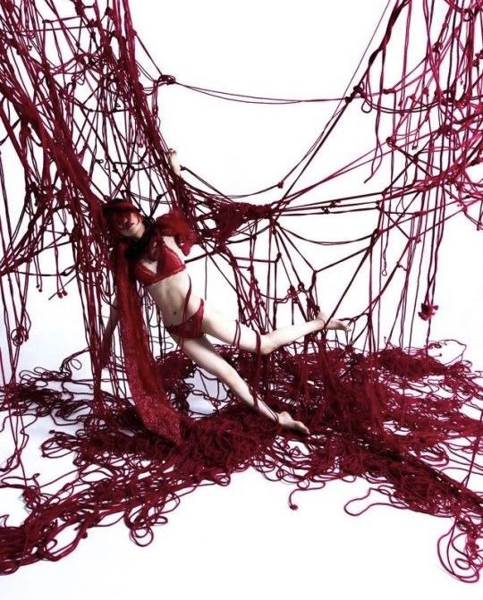the image shows a slender woman in red lingerie partially obscured and entangled within a massive, tangled web of thick, crimson string or rope. the string forms a complex, irregular network that extends throughout the frame.  the woman's pose suggests being trapped or held within the mass of red fibers. the overall mood is dark, artistic, and slightly sensual, hinting at themes of confinement or entrapment. the background is predominantly white, creating a stark contrast with the deep red of the ropes and the model's clothing. the photo is high quality and has a distinct artistic style.
