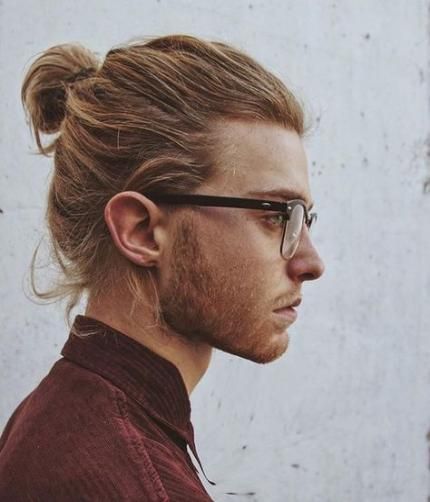 a profile shot of a man with blond hair styled in a bun, wearing glasses and a maroon shirt, with a beard.