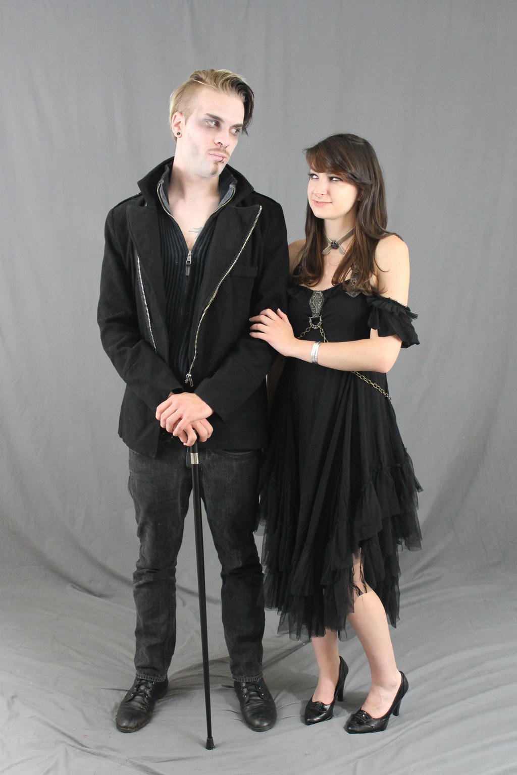 full shot of a couple dressed in gothic attire against a gray backdrop. the man, with pale makeup and short blonde hair, is wearing a black jacket, dark pants, and shoes, and carries a cane. the woman in a black dress with layers of tulle features a choker necklace and her dark brown hair is styled down. they stand close, the woman's arm around the man's, creating a romantic and mysterious mood.  the setting is clearly a studio, with a neutral, even lighting.