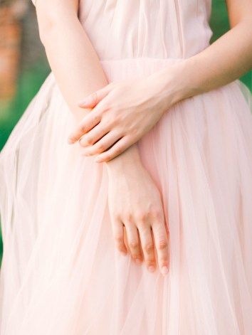 closeup view of a woman's hands gently resting on her light pink tulle dress.  the dress is flowing and elegant, and the overall aesthetic is romantic and soft. the background is blurred, suggesting an outdoor setting. the focus is on the woman's hands and the texture of the dress.