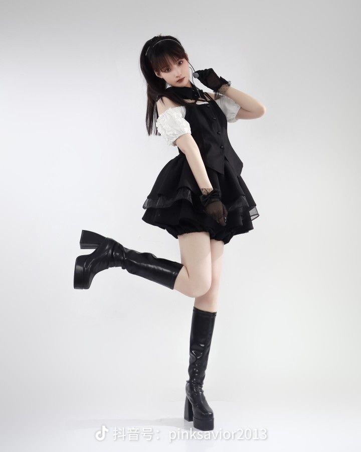 a young woman in a gothicinspired outfit poses in a studio. she's wearing a black and white dress with a layered skirt, fingerless gloves, and platform boots. she has dark hair styled with a headband. her right leg is raised. she is looking at the camera. the background is a plain white.