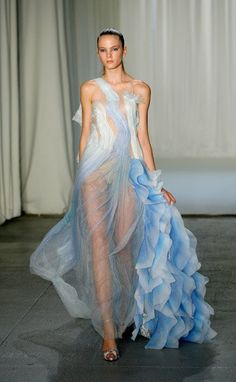 a woman in a white and blue tulle gown poses on a runway. the dress is sheer and has a flowy, ethereal look. the model is wearing her hair up and has a natural makeup look. the background is a white backdrop, with a few glimpses of a curtain in the background.